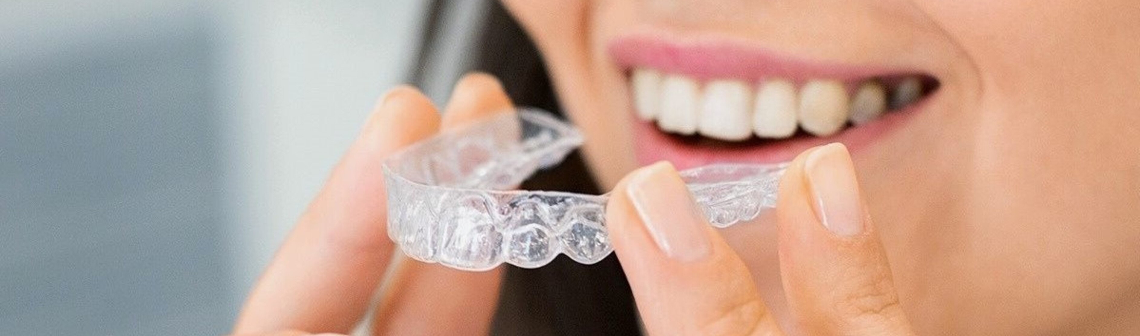 Donna sorridente con denti bianchi tiene tra le dita un allineatore dentale trasparente in plastica, su sfondo chiaro