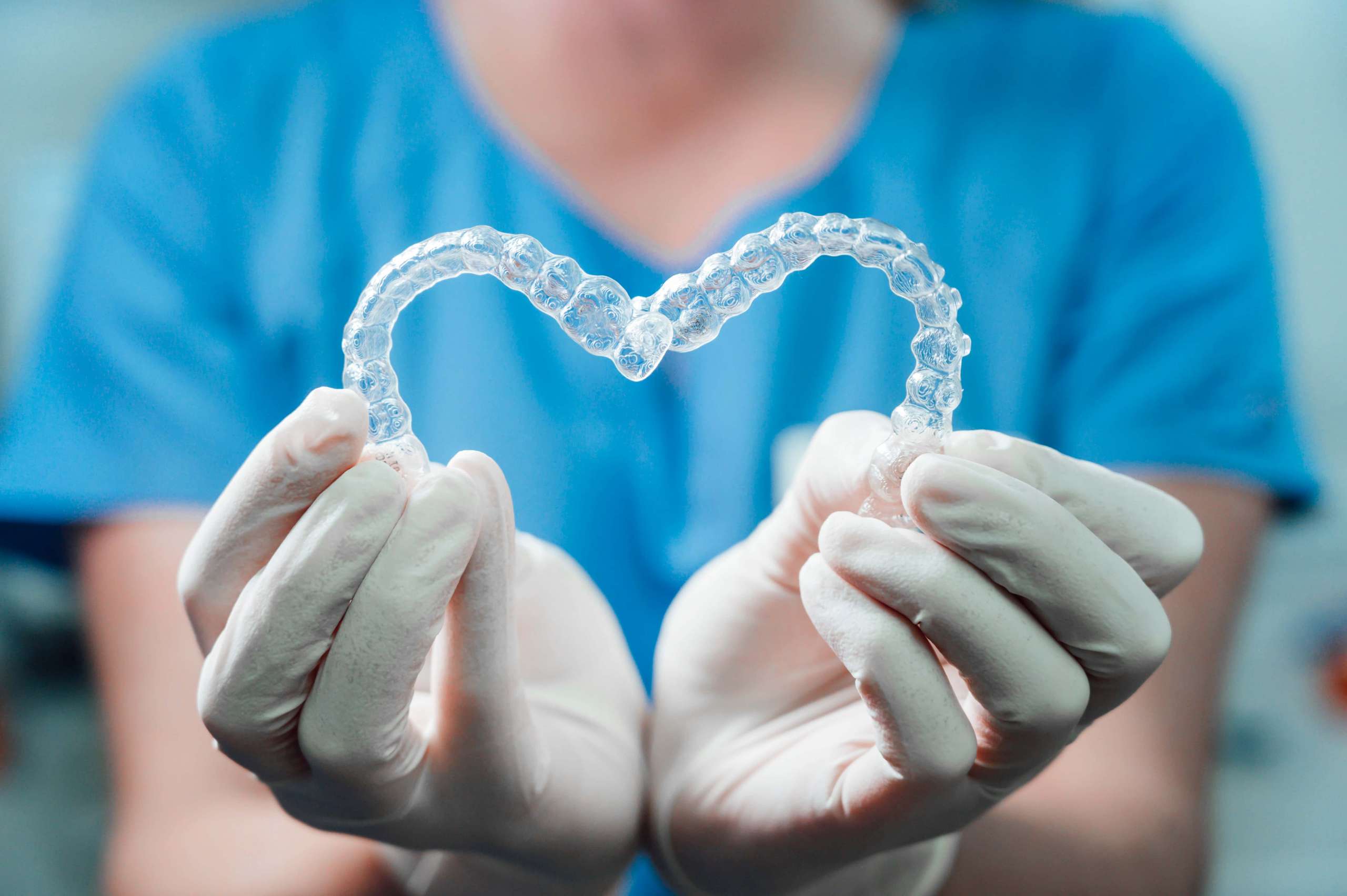 Mani in guanti bianchi piegano un allineatore dentale trasparente a forma di cuore davanti a una divisa azzurra